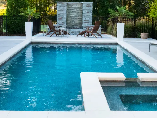 A backyard pool with clear water and a shallow wading area separated by a mosaic tile border. The pool steps and water surface reflect the sky.
