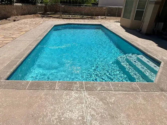 A backyard with a small rectangular pool and an attached square hot tub, both empty. The pool is surrounded by tiled pavement and stone walls, with houses visible over the wall. Pool floats and garden chairs are stacked nearby.