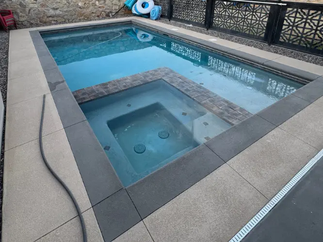 A backyard with a small rectangular pool and an attached square hot tub, both empty. The pool is surrounded by tiled pavement and stone walls, with houses visible over the wall. Pool floats and garden chairs are stacked nearby.