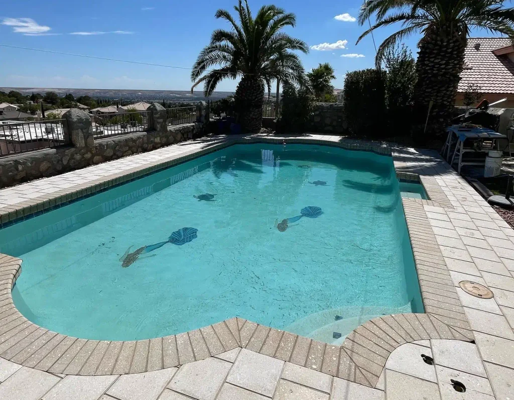A backyard with a small rectangular pool and an attached square hot tub, both empty. The pool is surrounded by tiled pavement and stone walls, with houses visible over the wall. Pool floats and garden chairs are stacked nearby. Understanding the Role of a Pool Contractor Blog